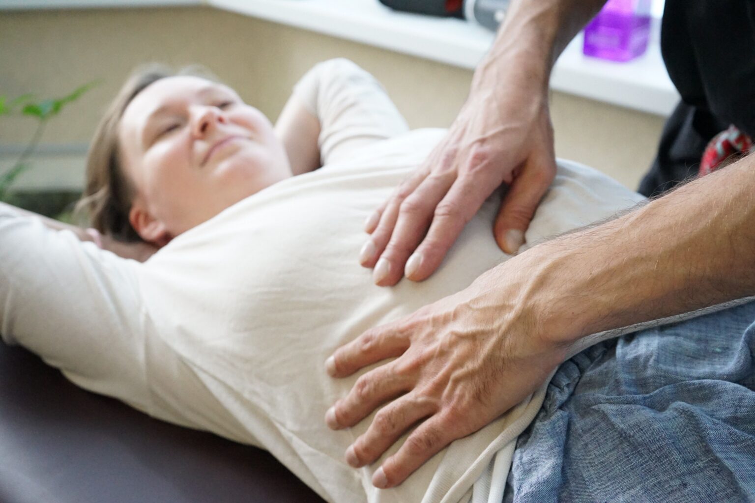 Mains d'un ostéopathe posées sur l'abdomen d'une patiente allongée, bras relevés
